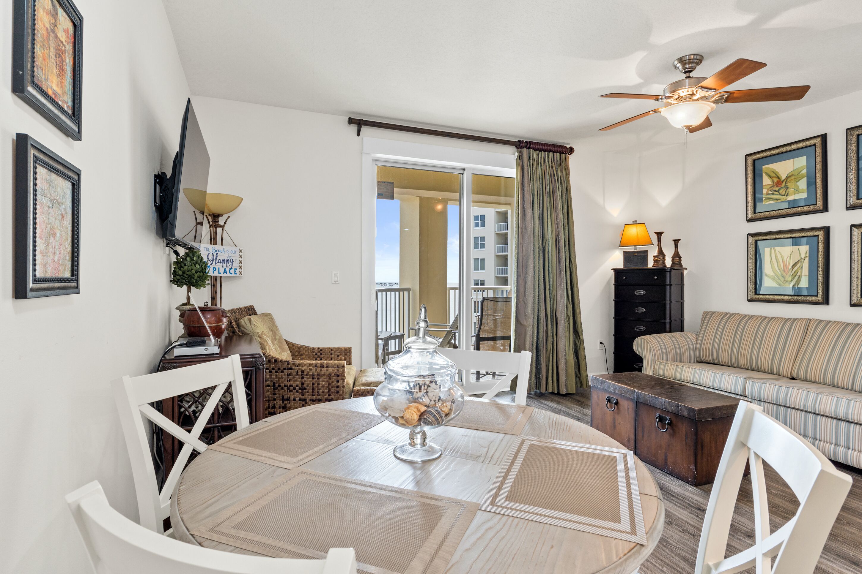 Un coin repas avec une table ronde, des chaises, un ventilateur de plafond et une vue sur un balcon extérieur, accessible par une porte vitrée.