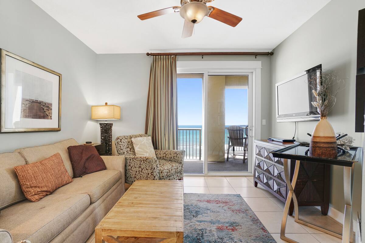 A living room with a sofa, coffee table, and a TV on a stand. There is a balcony with a view of the sea.