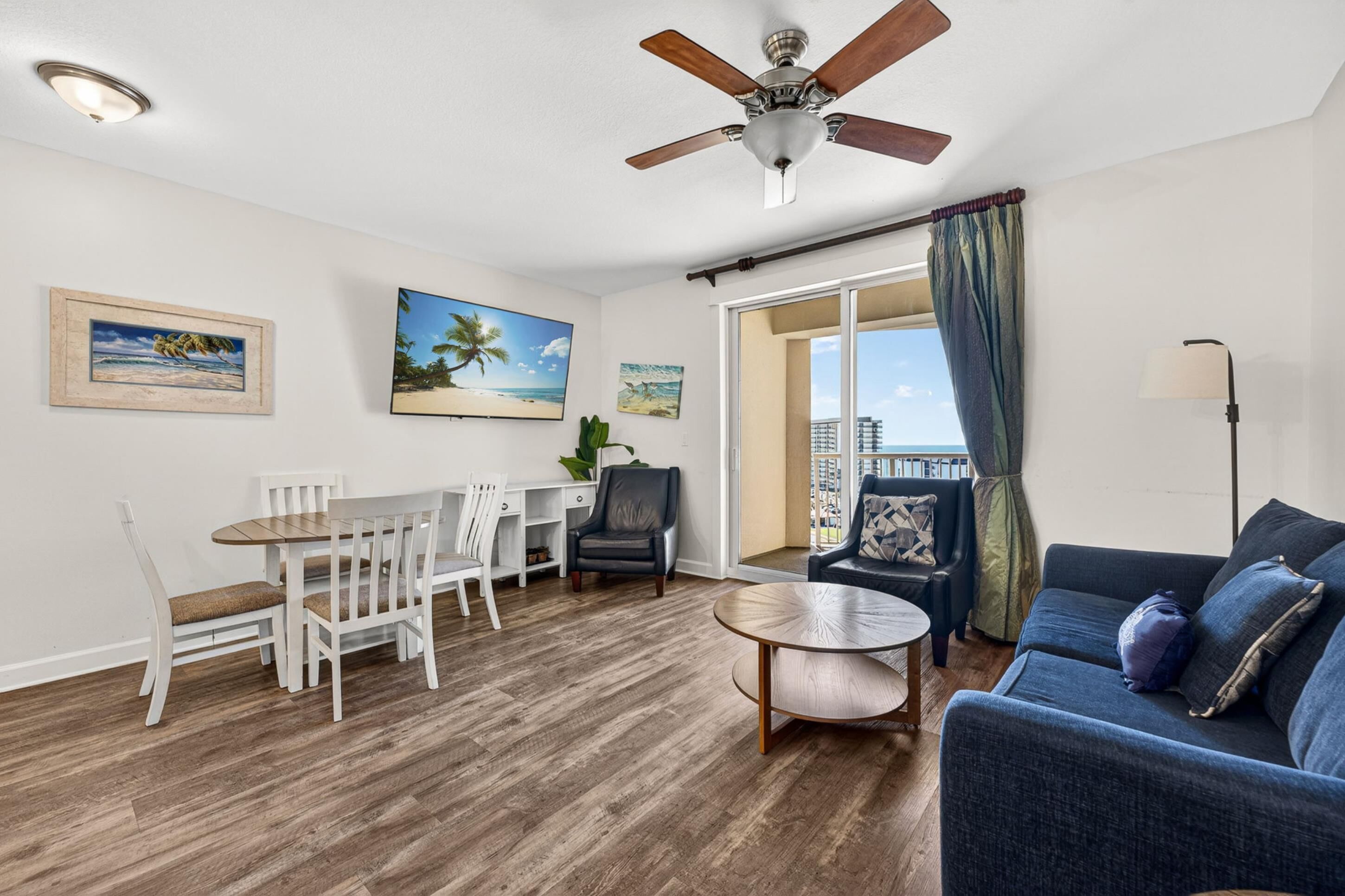 Ein Zimmer mit Deckenventilator, Esstisch mit Stühlen, einer Couch und Blick ins Freie.