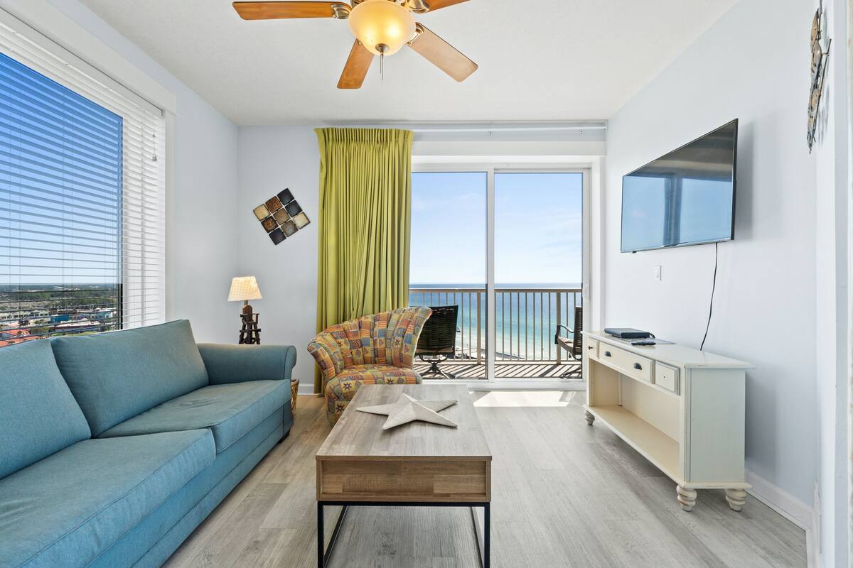 A modern living room with a blue sofa, a wooden coffee table, a colorful armchair, a green curtain, a ceiling fan, a TV, and a balcony with a view of the ocean.