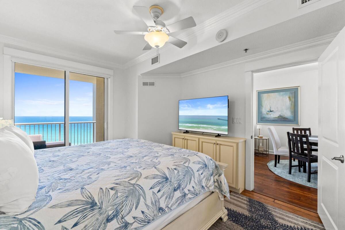 A bedroom with a bed, a television, a painting, and a balcony with a view of the ocean.