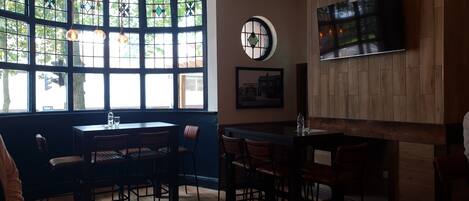 A restaurant with a curved wooden counter, multiple tables, and chairs. There are large windows with decorative glass panes, a round wall clock, and a flat-screen TV mounted on the wall.
