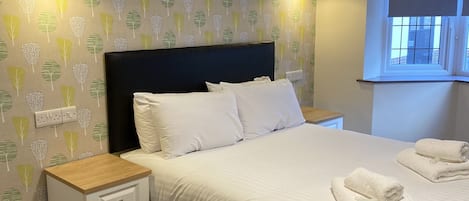 A neatly made bed with white linens and two towels folded on top, a bedside table with a drawer, a wall-mounted lamp, and a window with a blind.