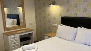 A neatly made bed with white linens and two folded towels, a wooden dresser with drawers, a small bedside table with a lamp, and a wall with a leafy pattern. - The Bournbrook Inn (Birmingham)