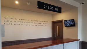 A reception area with a wooden counter, a 'CHECK IN' sign, a mounted television, and a wall with a quote about inns. - The Bournbrook Inn (Birmingham)
