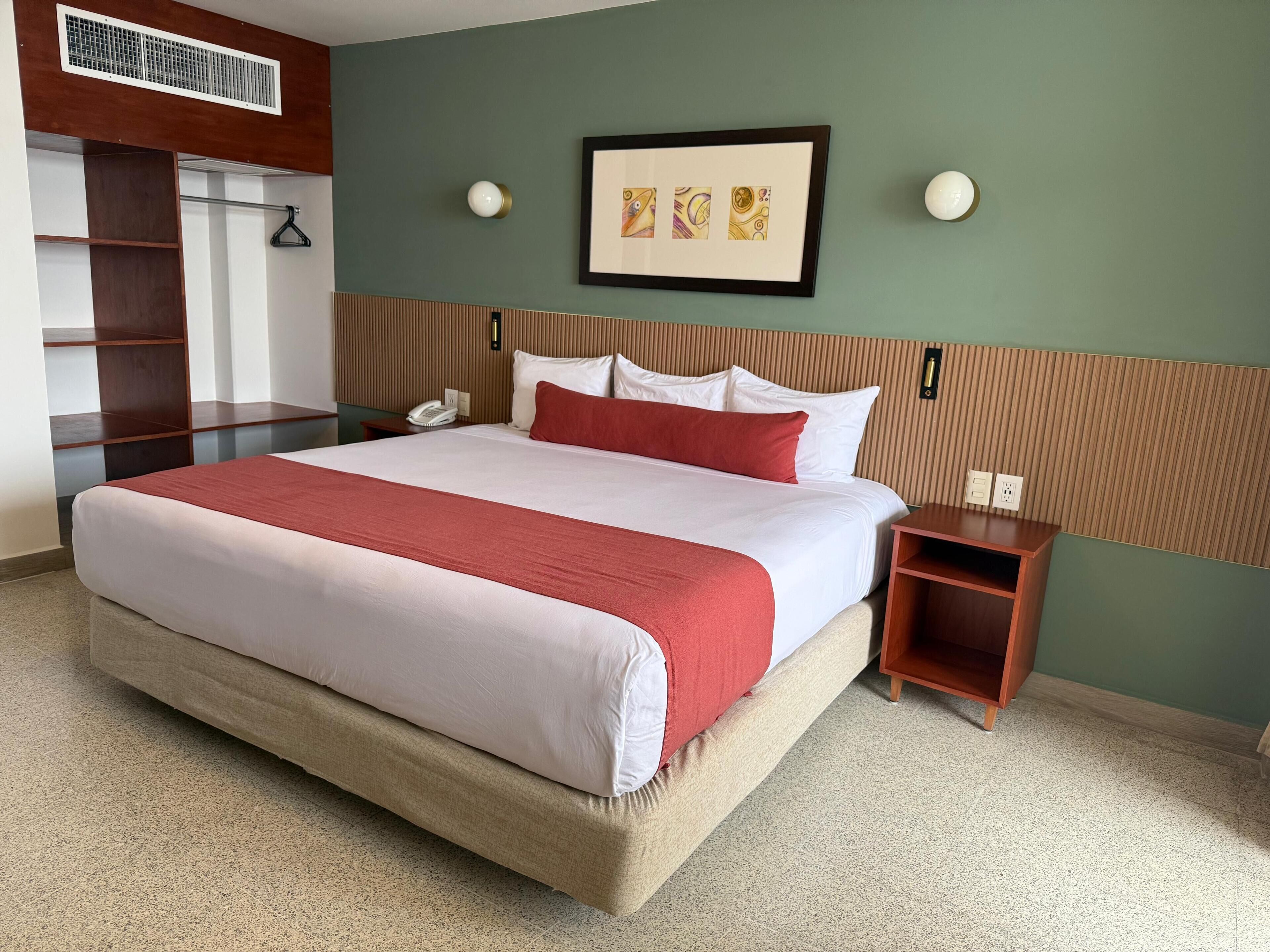 A hotel room with a large bed, a wooden headboard, a bedside table with a phone, a framed artwork on the wall, and a coat rack.