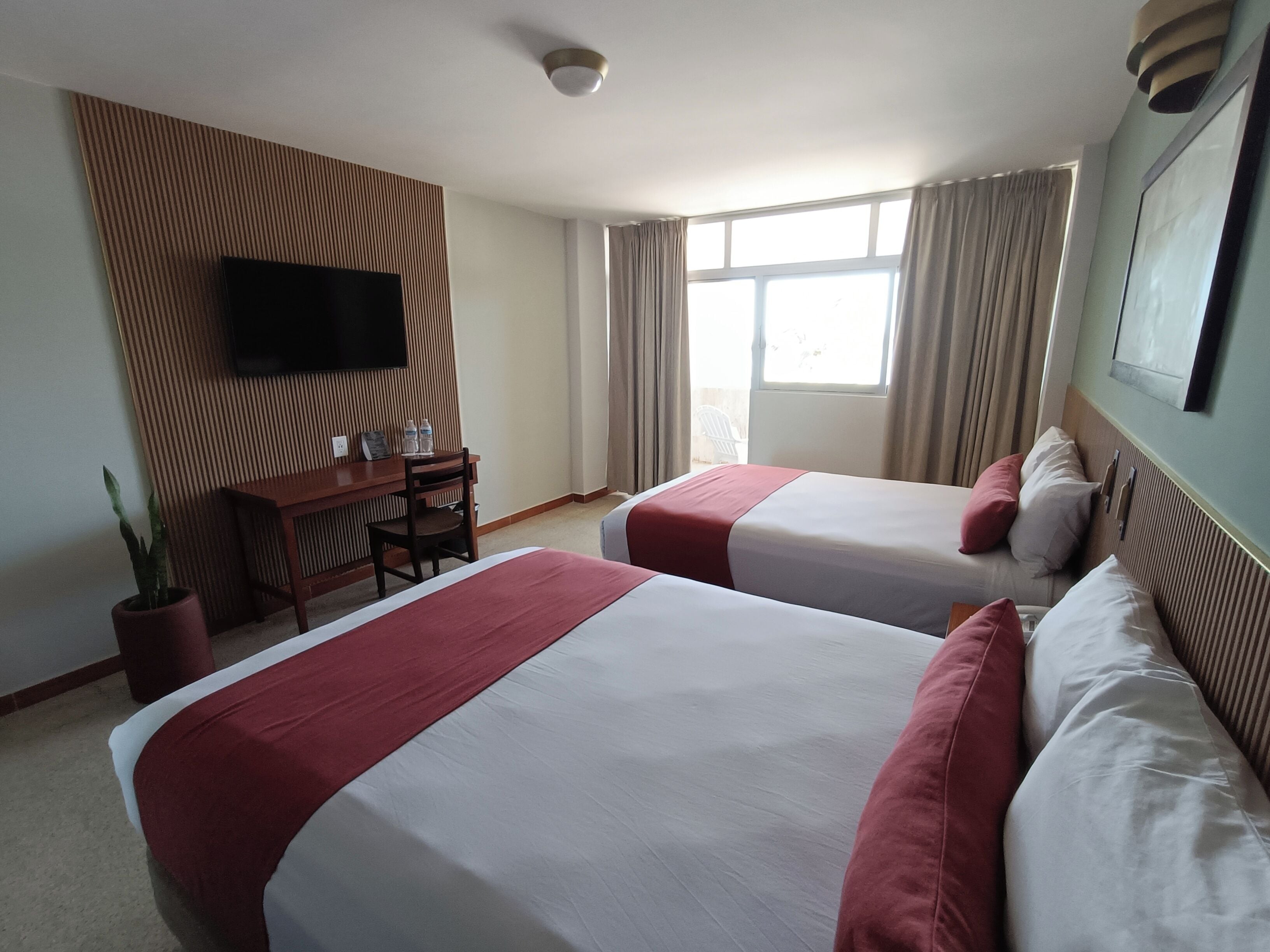 A hotel room with two beds, a desk with a chair, a television mounted on the wall, and a window with curtains.