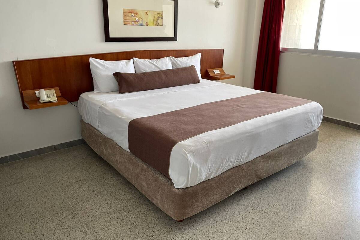 Une chambre d’hôtel avec un grand lit, une tête de lit en bois, une table de chevet avec un téléphone, un tableau encadré au mur et une fenêtre avec des rideaux.
