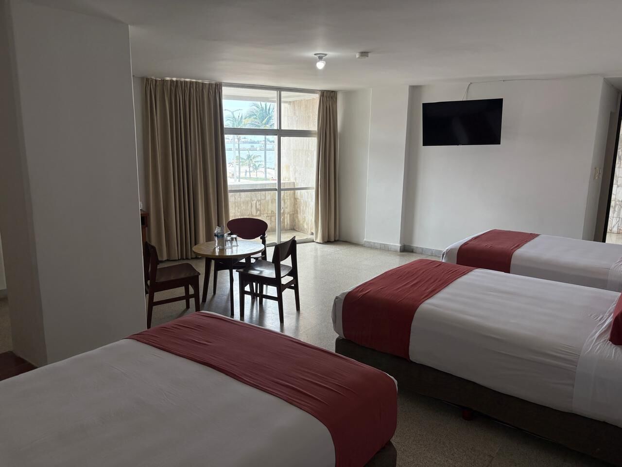 Une chambre d’hôtel avec trois lits, un bureau avec des chaises et une grande fenêtre avec des rideaux.
