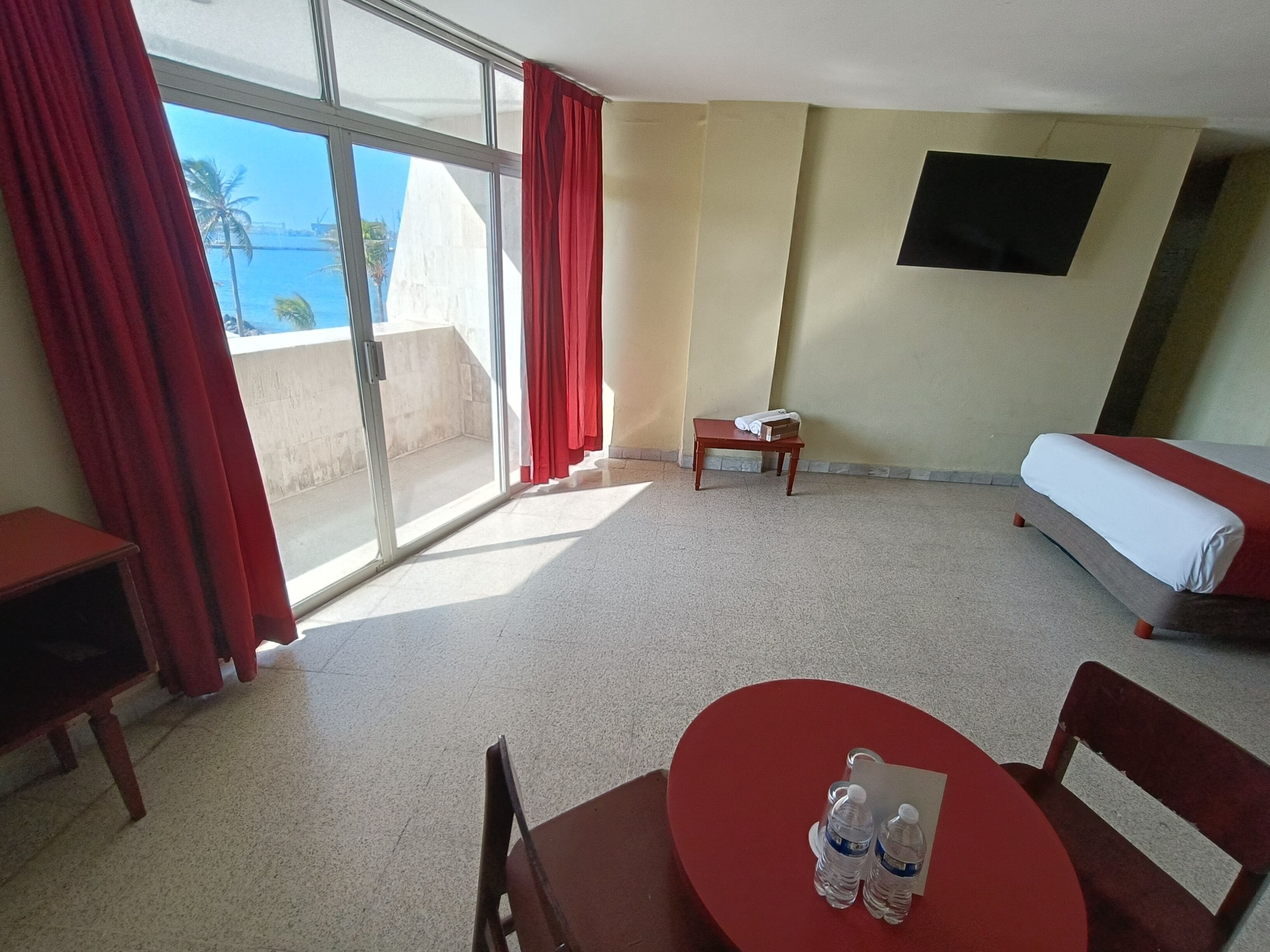 A hotel room with a bed, a small table with water bottles, a TV on the wall, and a view of the sea through large windows.