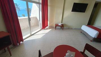 A hotel room with a bed, a small table with water bottles, a TV on the wall, and a view of the sea through large windows.