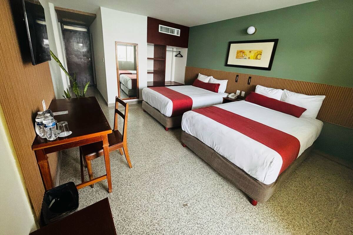 Une chambre d’hôtel avec deux lits, un bureau, une chaise, un miroir, un téléphone fixé au mur et un tableau encadré.