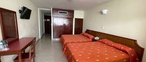 Habitación de hotel con dos camas individuales, un escritorio con silla, televisor montado en la pared y una puerta que da a un baño.