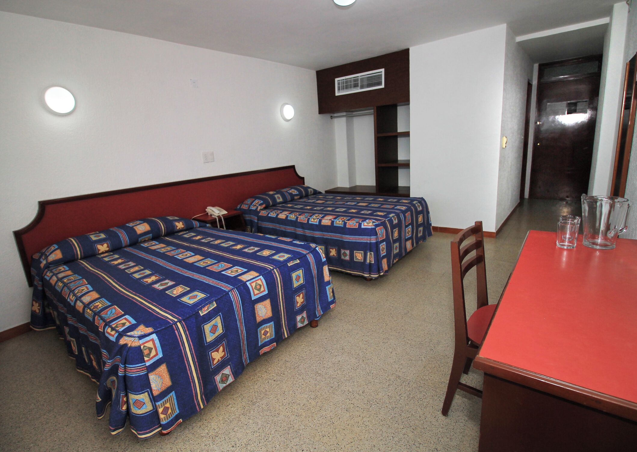 A hotel room with two beds, a red table, and a small kitchenette.