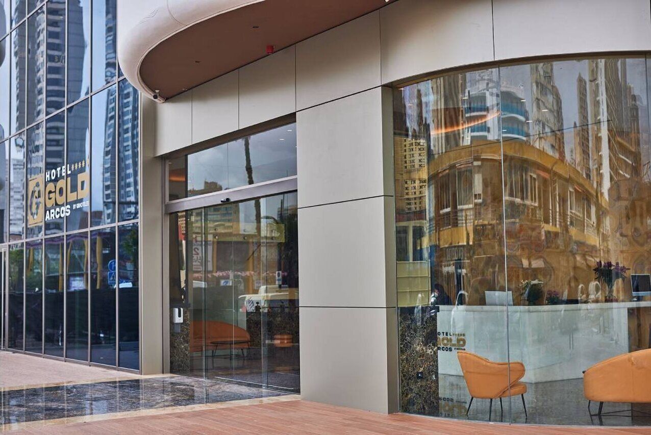 A hotel with a glass facade, a reception desk, and orange chairs outside.