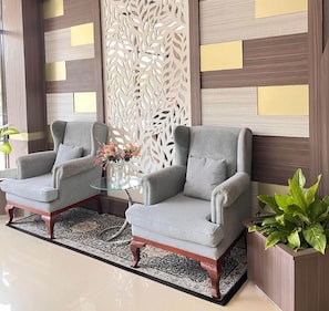 Una habitación con dos sillones grises, una mesa de centro de cristal con flores, una alfombra estampada y un panel blanco decorativo.
