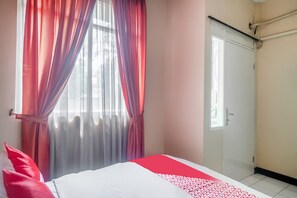 Habitació d'hotel amb un llit, roba de llit vermella i blanca, una finestra amb cortines de tul i una porta.