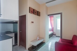 Espai habitable petit amb cuina americana, un sofà vermell, una petita banca i una porta que condueix a un dormitori amb llit i cortines.