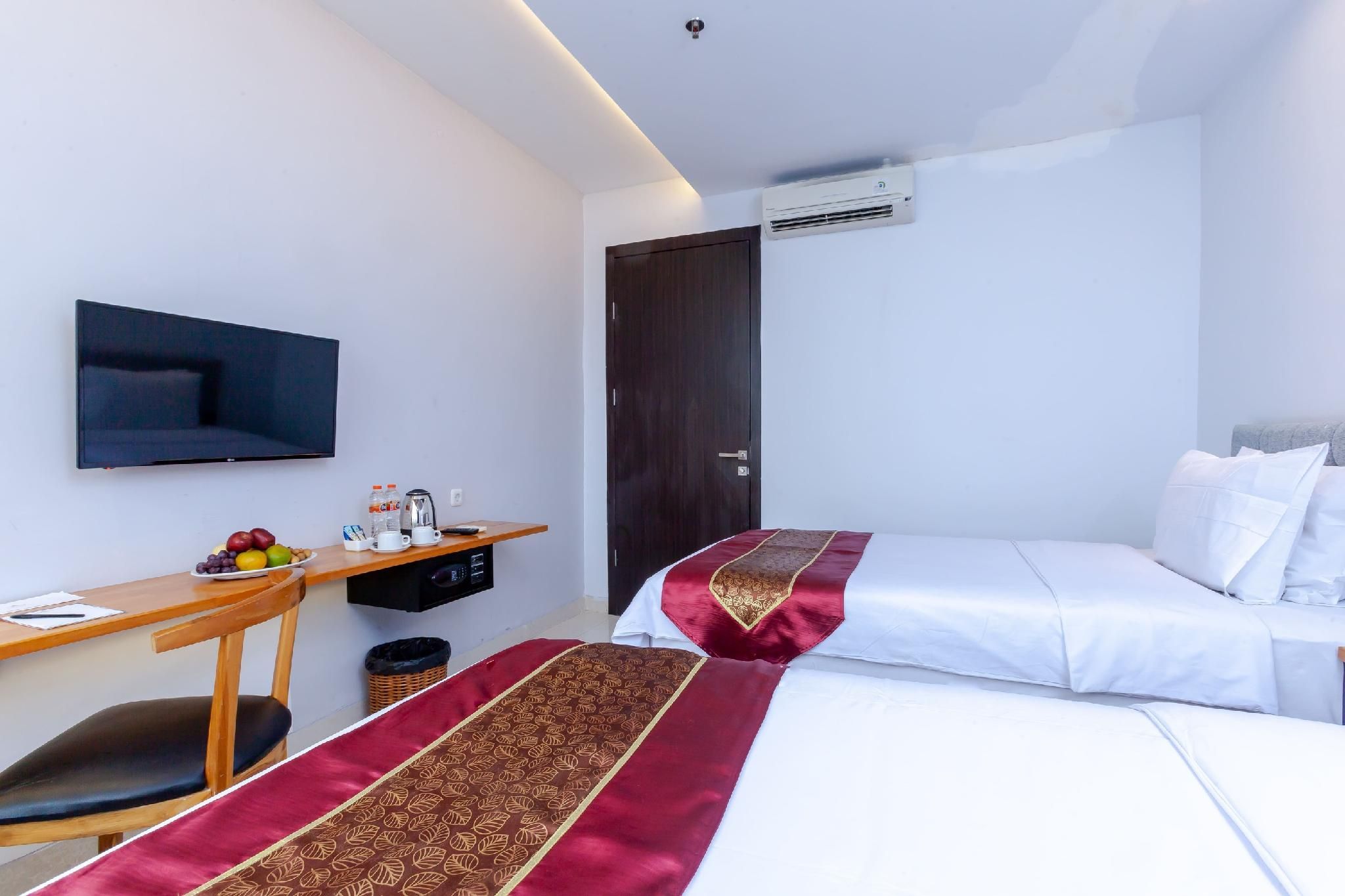 Habitación de hotel con cama, escritorio con televisor de pantalla plana, mesita con fruta y cafetera, y unidad de aire acondicionado en la pared.