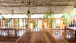 A spacious indoor area with a wooden table and benches, a thatched roof, and potted plants.