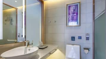 A modern bathroom with a large oval sink, a mirror, a framed artwork on the wall, and a toilet with a towel dispenser.
