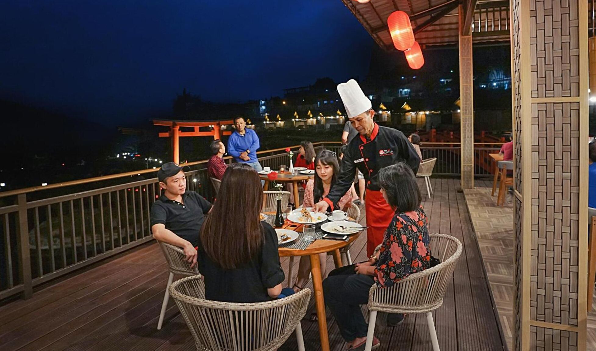 A group of people dining at an outdoor restaurant with wicker chairs and a wooden deck.