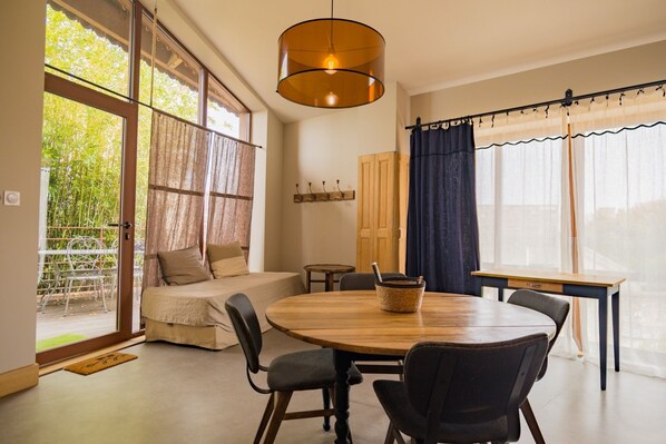 Un comedor con una mesa redonda de madera, sillas, una lámpara colgante, un sofá y vistas al exterior con vegetación.