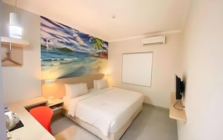 Habitación de hotel con cama, escritorio, silla, un cuadro de una playa y televisor sobre un soporte.