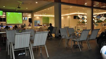 Ein moderner Innenbereich mit Sitzgelegenheiten, Tischen und Stühlen, einem großen Bildschirm, der ein Sportereignis zeigt, und einer Glastrennwand, die den Sitzbereich von der Bar oder Lounge abgrenzt.