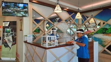Un espace d’accueil d’hôtel comprenant un comptoir, un téléviseur fixé au mur et une grande fenêtre donnant sur une plage.
