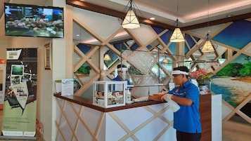 Zona de recepción de un hotel con mostrador, televisor montado en la pared y un ventanal con vista a la playa.