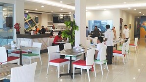Un moderno comedor con sillas y mesas blancas, una barra con personal y un amplio ventanal que proporciona luz natural.