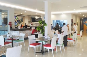 Un moderno comedor con sillas y mesas blancas, una barra con personal y un amplio ventanal que proporciona luz natural.