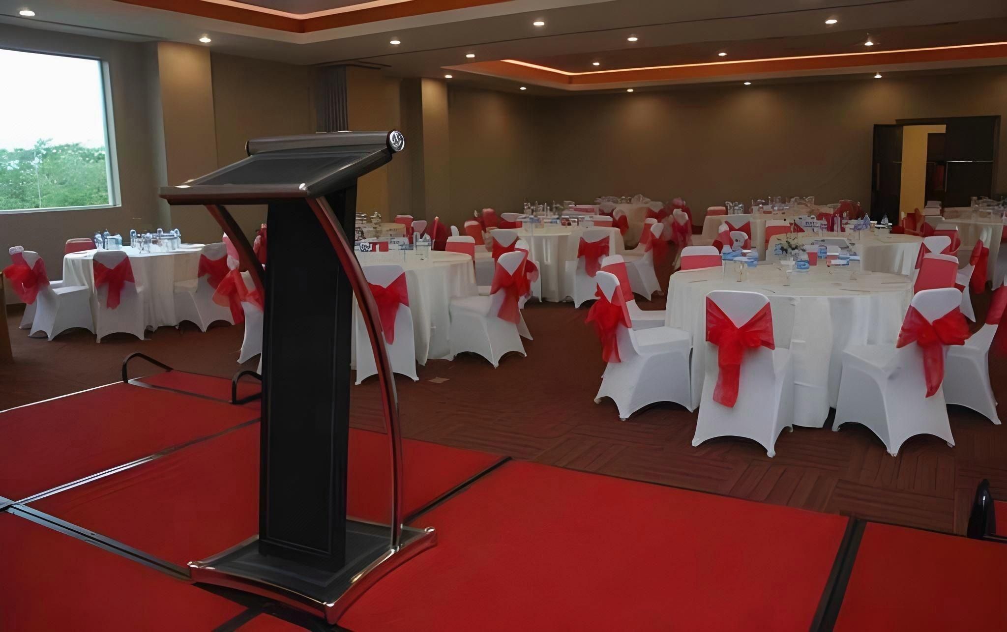 A conference room with multiple round tables, each with white tablecloths and red sashes, and a podium in the foreground.