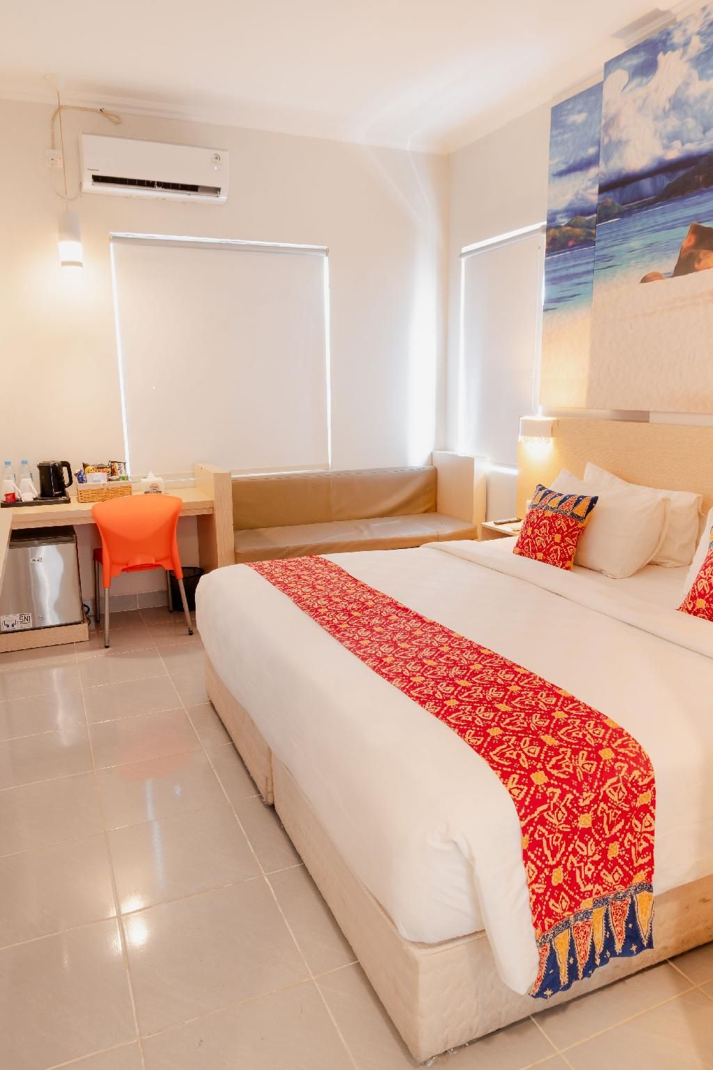 A hotel room with a bed, a desk, a chair, a small kitchenette, and a painting on the wall.