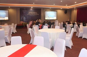 A conference room with round tables and white chairs, a stage with a presentation screen, and a banner with text. - Cordela Hotel Pangkal Pinang (Pangkalpinang)
