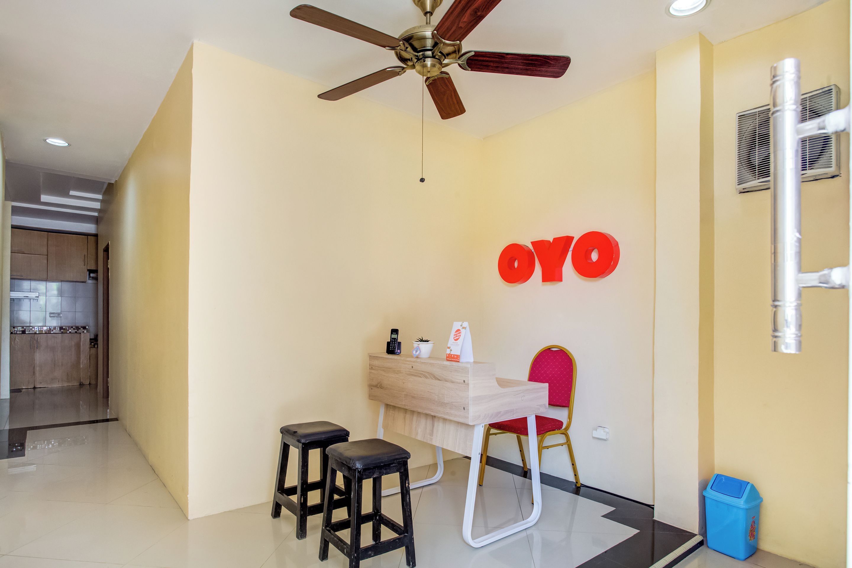A hotel room with a wooden desk, two stools, a red chair, a ceiling fan, and a wall-mounted air conditioning unit.