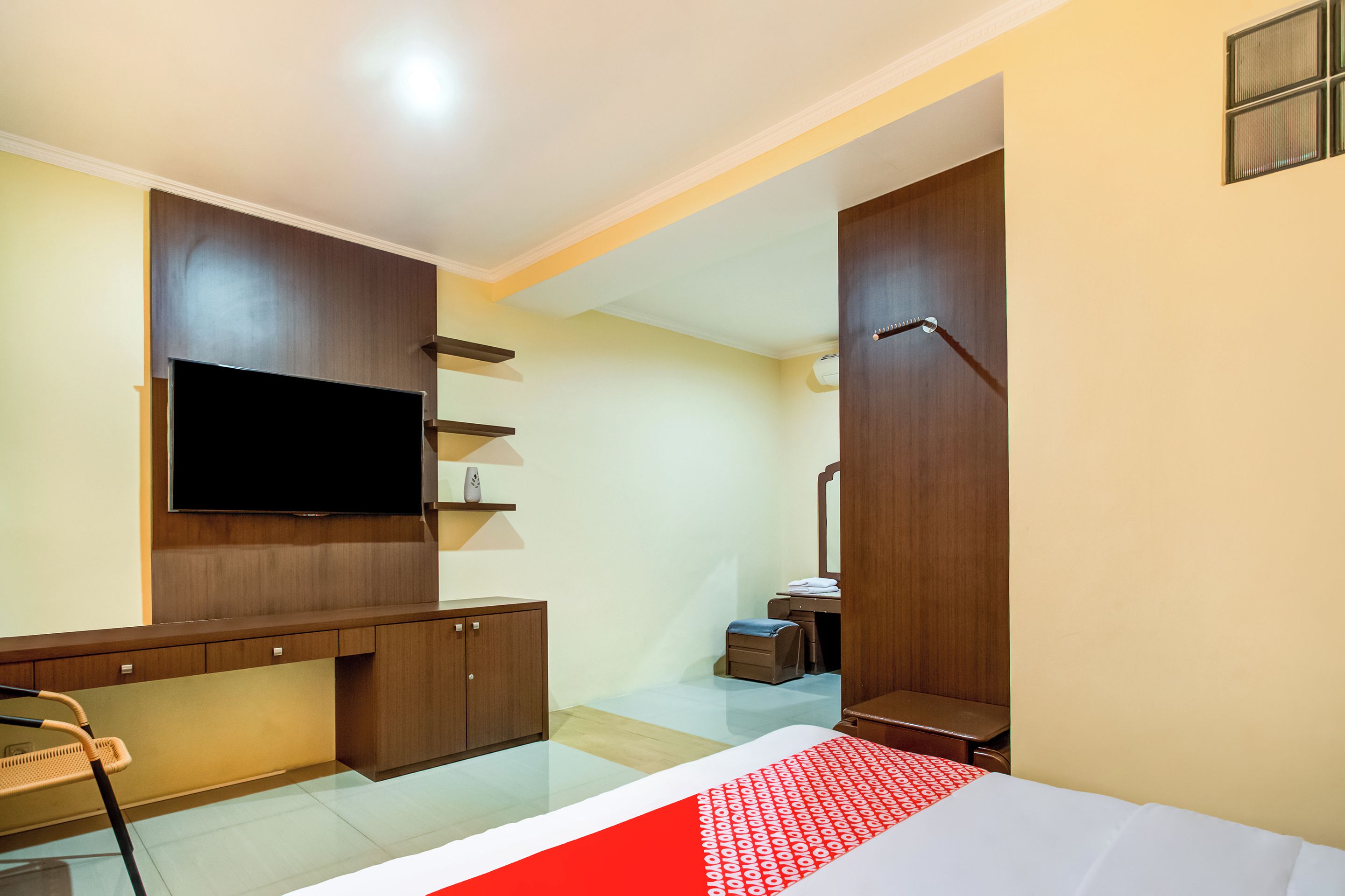 A hotel room with a flat-screen TV mounted on the wall, a wooden cabinet, a small table with a chair, and a bed with a red and white patterned bedspread.