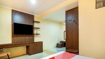 A hotel room with a flat-screen TV mounted on the wall, a wooden cabinet, a small table with a chair, and a bed with a red and white patterned bedspread.