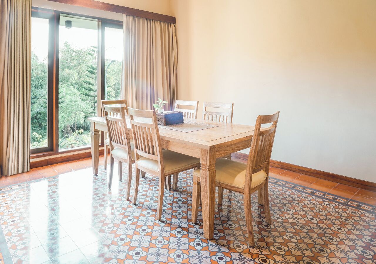 A dining area with a wooden table and chairs, large windows with curtains, and patterned floor tiles.