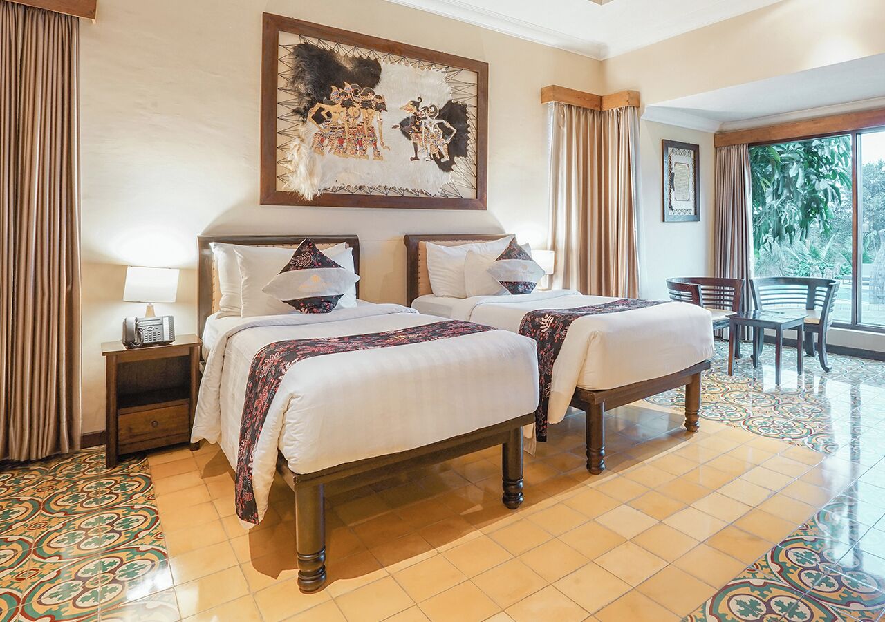 A hotel room with two beds, a large window, and a view of greenery outside.