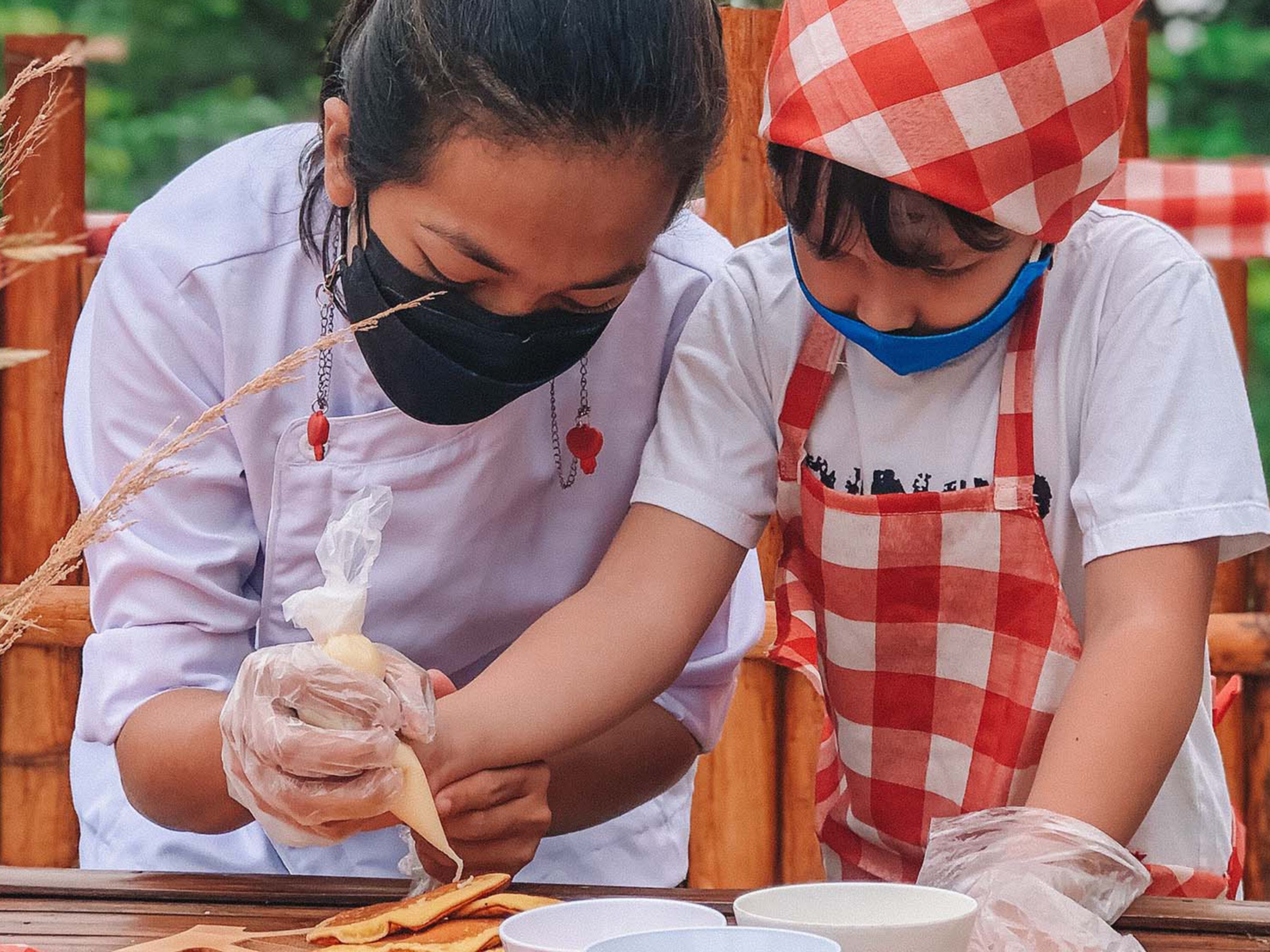 Two individuals preparing food, one wearing a checkered apron and a hat, and the other in a white shirt and black mask.