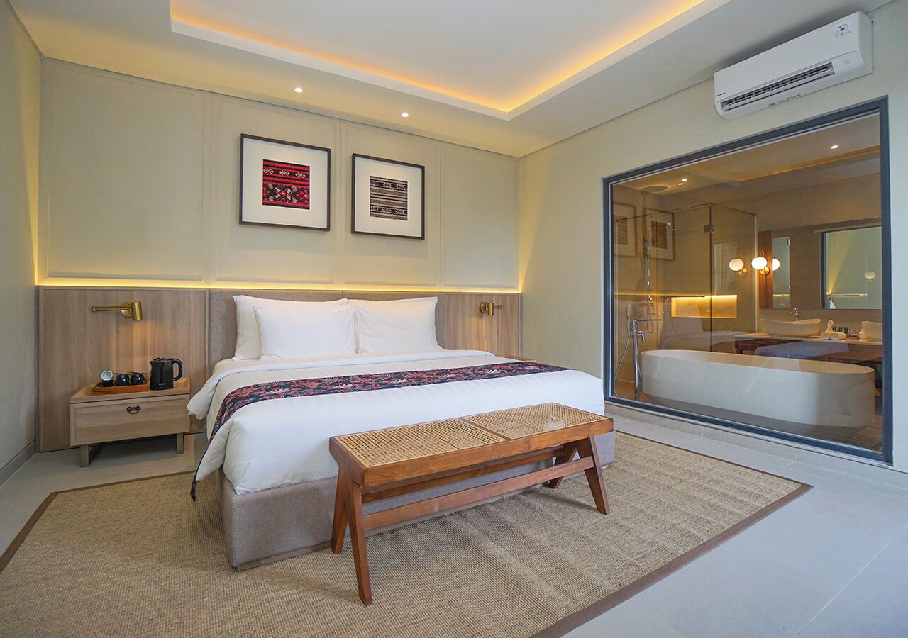 A modern hotel room with a large bed, bedside tables, a bench, and a glass-enclosed bathroom.