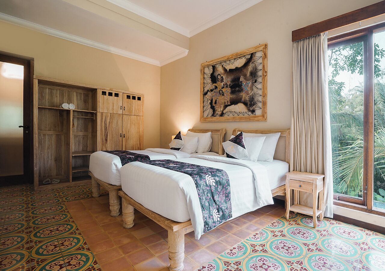 A bedroom with two beds, a wooden wardrobe, a large painting, and a view of greenery through a window.