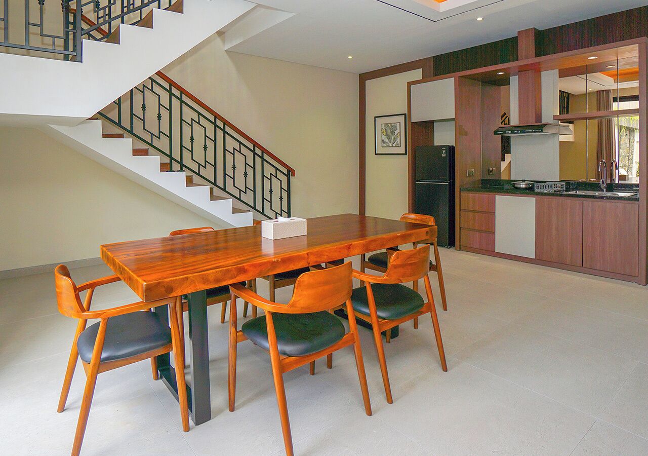 A dining area with a wooden table and chairs, a staircase, and a kitchen area with a refrigerator and stove.