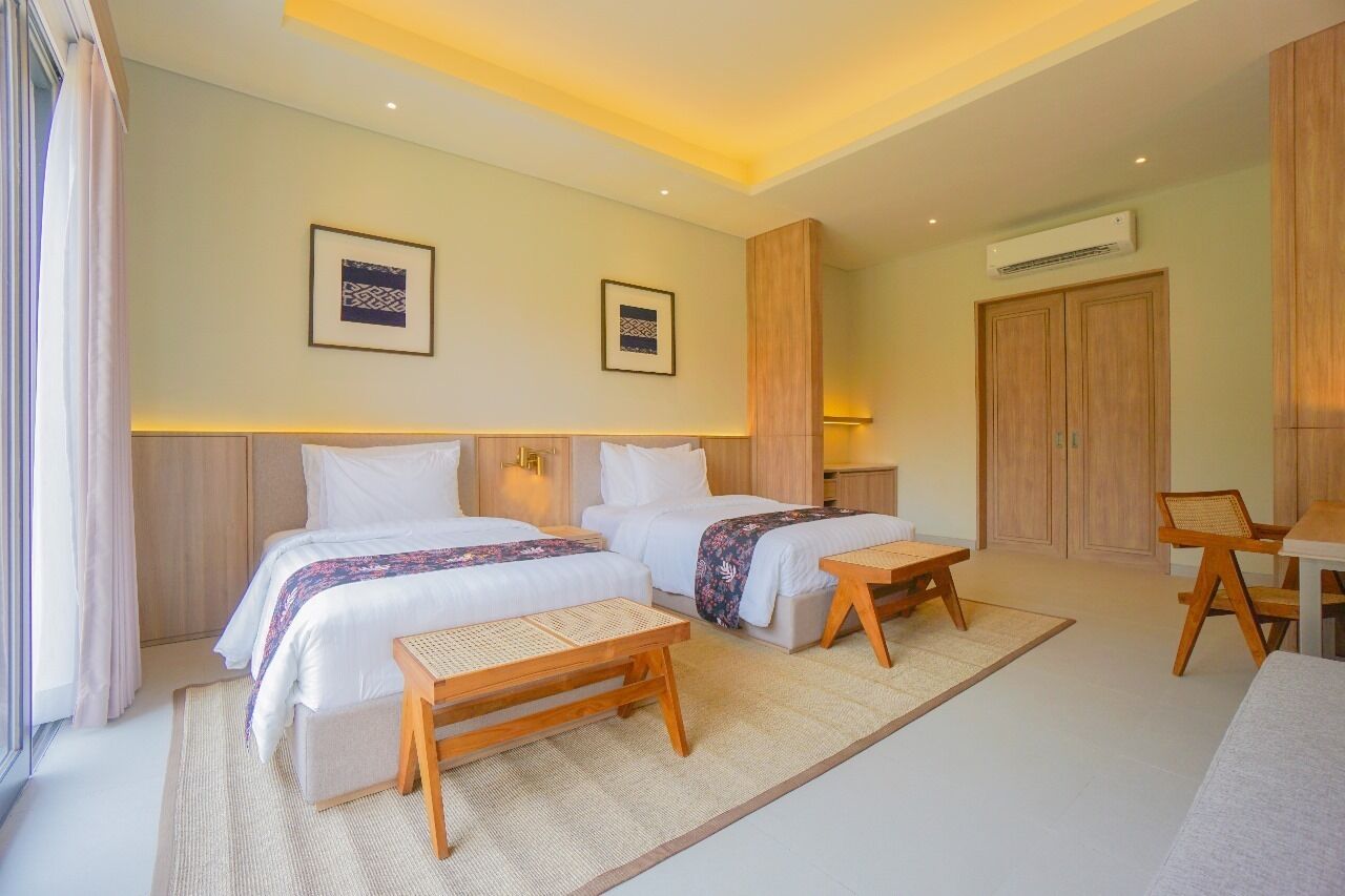 Habitación de hotel con dos camas, un escritorio y una silla. Se observan dos cuadros en la pared, un armario de madera y un equipo de aire acondicionado.