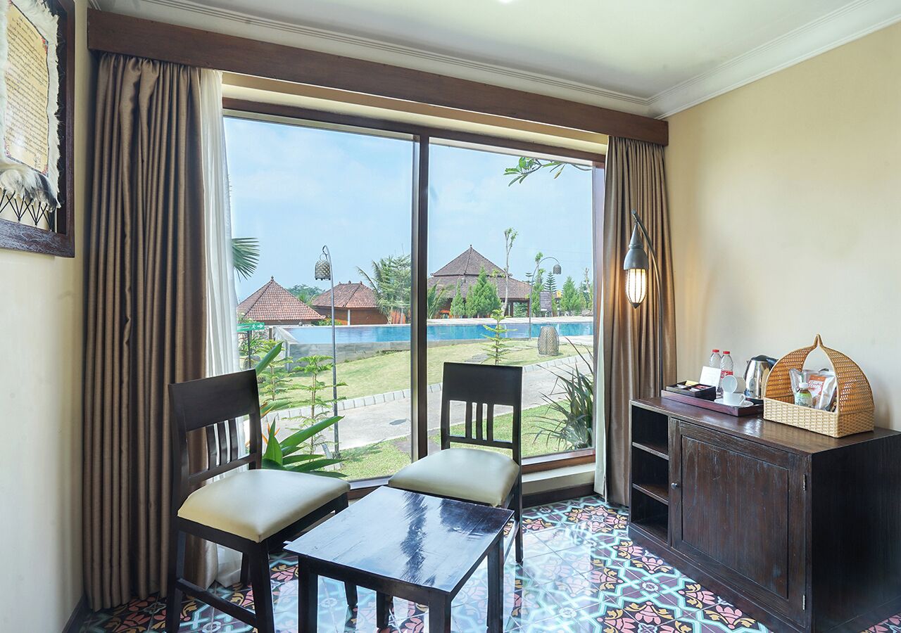 A room with two chairs, a small table, a cabinet, and a view of a swimming pool and buildings outside.