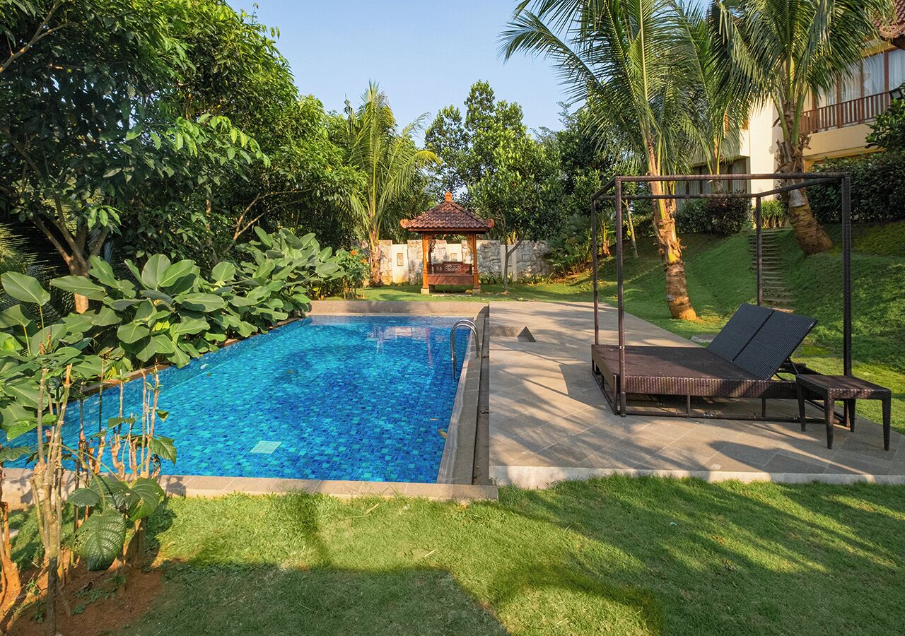 Una piscina con solárium de baldosas, rodeada de abundante vegetación y un pabellón.