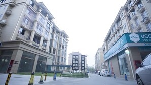 Un edificio moderno de varios pisos, con amplias ventanas y balcones. Varias coches están estacionados adelante, y un letrero con caracteres chinos.