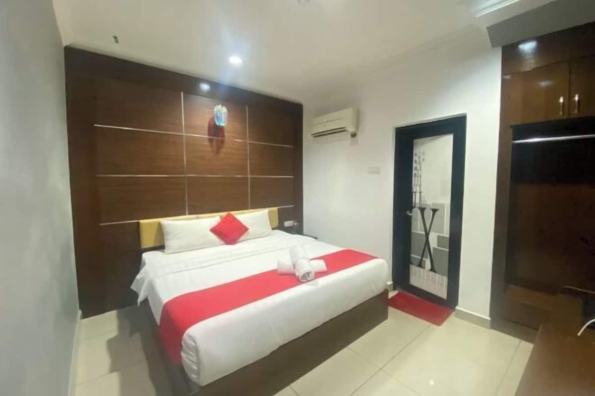 Una camera d'hotel con un letto, una testiera in legno, un condizionatore d'aria, una porta con uno specchio e una moquette rossa.